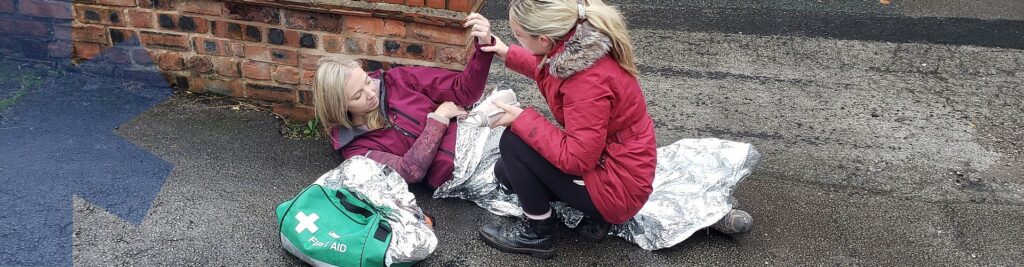 Picture of a blonde haired lady in a red coat providing first aid to another blonde haired lady who is lying on the floor with a silver blanket over them.