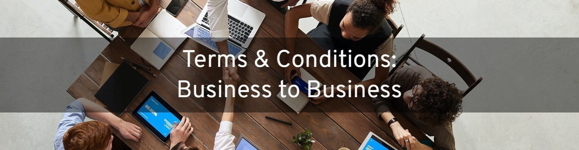 Terms and Conditions (Business to Business). Image shows these words over a picture of business people sitting around a wooden table with two people on opposite sides shaking hands.